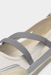 Sandale blanche et beige avec deux larges bandes élastiques grises sur le dessus et une semelle basse et plate.