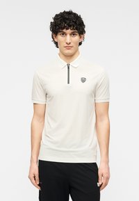 Μπλούζα πόλο σε ανοικτό κρεμ χρώμα, με κοντά μανίκια, διαθέτει φερμουάρ στην τρύπα και μαύρο λογότυπο στο αριστερό μέρος του στήθους. Απαλή, ομαλή υφή.