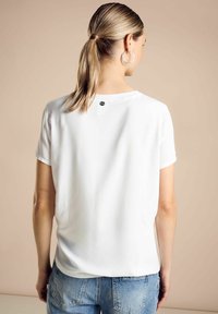 Blusa bianca a maniche corte con scollatura rotonda e leggero lucido. Presenta un piccolo etichetta nera sul retro vicino alla scollatura. Abbinata a jeans blu.