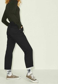 Person klädd i svart långärmad topp och svarta byxor, med händerna i fickorna, samt vita och svarta tie-dye strumpor och bruna låga sneakers.