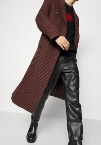 Dubbelknäppt kappa i ett röd och svart houndstooth-mönster, tillverkad av texturerad tyg. Matchad med svarta läderbyxor och ankle boots.