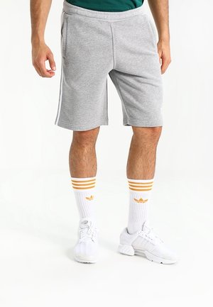 Man draagt grijze sportshorts, witte sneakers en witte crew sokken met gele strepen en logo's op een witte achtergrond.