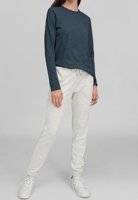 Kvinna i mörk teal långärmad skjorta, ljusgrå joggingsbyxor med snörning i midjan och vita sneakers som står mot en grå bakgrund.