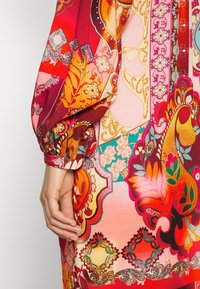 Värikäs kuviollinen mekko, joka sisältää sekoituksen punaisia, oransseja ja vaaleanpunaisia sävyjä. Pitkät puffihihat, joissa on joustavat hihansuut. Monimutkaisia kukka- ja geometristen kuvioiden suunnitteluja.