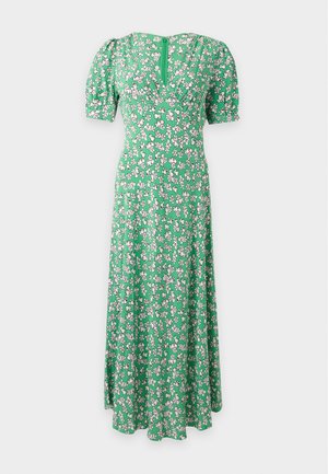 Vrouw staat en draagt een groene midi jurk met V-hals, een witte en roze bloemenprint en korte pofmouwen, gecombineerd met lichtbeige sandalen met hoge hakken.
