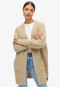 Beige strikket cardigan med åben front, ribbet tekstur, to for- lommer og lange ærmer, parret med en hvid skjorte og blå jeans.
