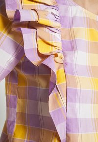Détail de manche à volants sur un haut en tissu à carreaux violet, jaune et beige avec une couture froncée près de la couture de l'épaule.