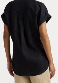 Black linen shirt with a curved hem, short rolled sleeves, and a center back pleat, shown from the back against a neutral background.
