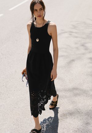 Γυναίκα που περπατά στο πεζοδρόμιο φορώντας μαύρο φόρεμα χωρίς μανίκια με δαντελωτή τρύπα, μαύρα σανδάλια με τρουκς, κρεμαστή αλυσίδα, κρατώντας γυαλιά ηλίου στο ένα χέρι.