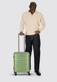 Uomo in maglione beige e jeans scuri in piedi con una mano in tasca, mentre tiene la maniglia di una valigia verde con ruote su uno sfondo grigio.