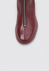 Bota de cuero burdeos hasta el tobillo con punta redonda, cremallera lateral y suela de goma texturada. Acabado suave con sutiles detalles de costura.
