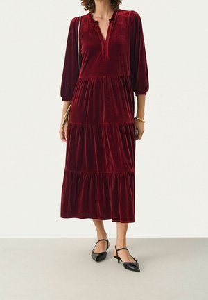 Kobieta w długiej bordowej welurowej sukience warstwowej z rękawami do łokcia i czarnych spiczastych butach na obcasie z paskiem.