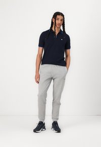 Polo marine avec un col zippé et des accents bleus, associé à un pantalon de survêtement gris chiné et des chaussures de sport foncées. Texture de tissu lisse.
