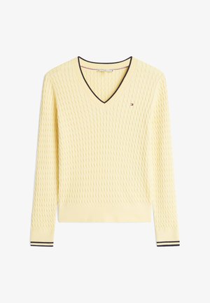 Dzeltena adīta džempera ar V veida izgriezumu, teksturētu dimanta rakstu un kontrastējošām melnām un baltām svītrām aprocēs. Mazs logotipa elements uz krūtīm.