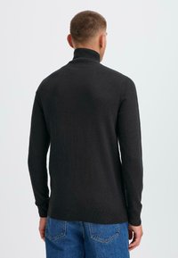 Svart stickad polotröja med långa ärmar och ribbad nederkant. Buren tillsammans med blå denimjeans, sedd bakifrån mot en ljus bakgrund.