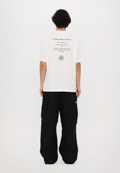 Λευκό μπλουζάκι με κείμενο στην πλάτη, μαύρο cargo παντελόνι, χαλαρή εφαρμογή, με τσέπες και λεπτομέρεια λογότυπου στο πλάι, ομαλή υφή υφάσματος.