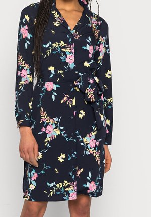 Femme portant une robe bleu marine à manches longues avec des motifs floraux roses, jaunes et bleus, boutonnée à l'avant, avec un nœud à la taille.