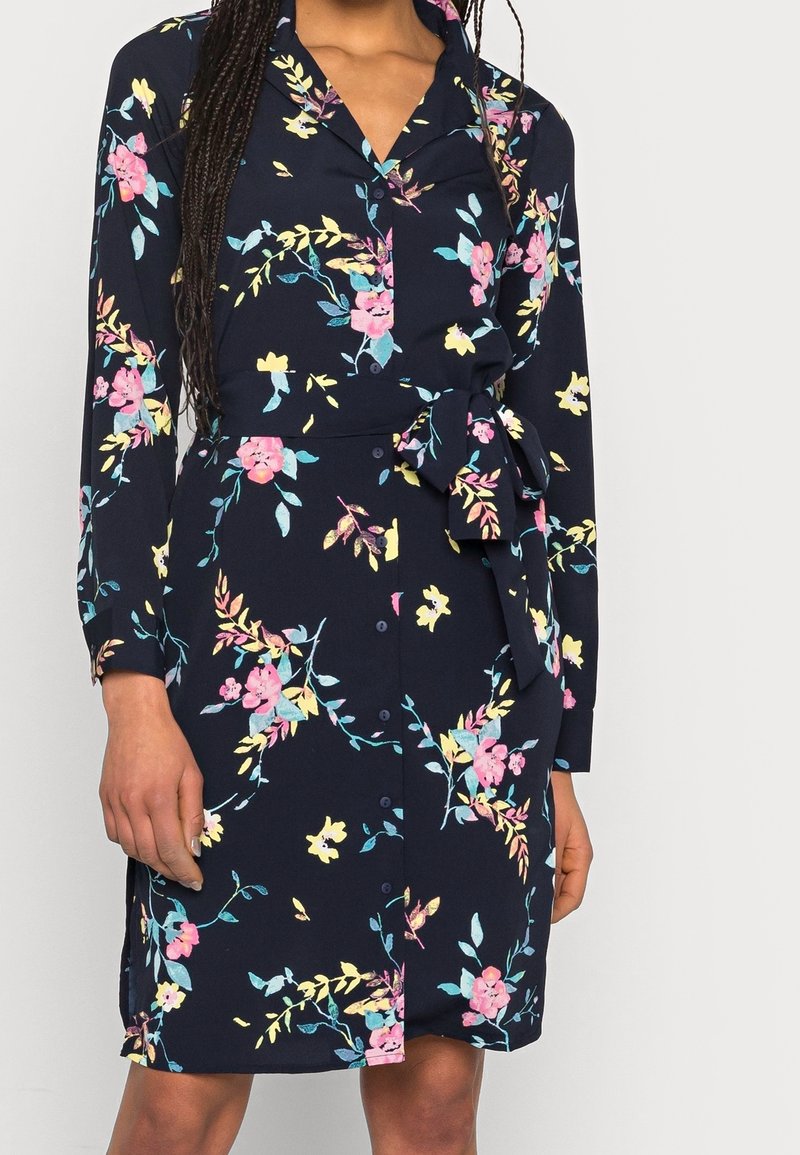 Femme portant une robe bleu marine à manches longues avec des motifs floraux roses, jaunes et bleus, boutonnée à l'avant, avec un nœud à la taille.