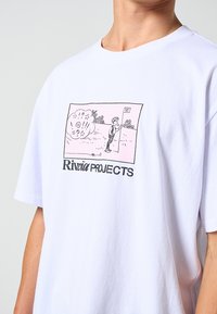 Λευκό βαμβακερό μπλουζάκι με ροζ γραφικό σχέδιο που απεικονίζει έναν χαρακτήρα που κρατά μια σημαία, με το κείμενο "Riviera Projects" σε μαύρο χρώμα κάτω από το σχέδιο.