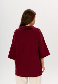 Camicia oversize borgogna con un ampio colletto, maniche corte e una texture liscia. La parte posteriore è semplice, senza motivi o embellishments visibili.