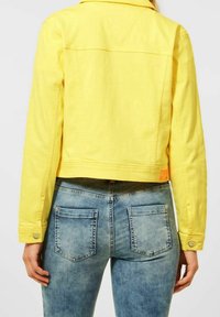 Gelbe Denimjacke mit einem verkürzten Design, geknöpften Manschetten und einem Rückenpasse-Detail, kombiniert mit blauen, ausgebleichten Denimjeans mit zwei Gesäßtaschen.