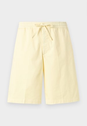 Άνδρας που στέκεται με τα χέρια στις τσέπες, φοράει λευκό πουκάμισο, λευκό μπλουζάκι, ανοιχτό κίτρινα σορτς με σκοινί και λευκά παπούτσια χωρίς κορδόνια.