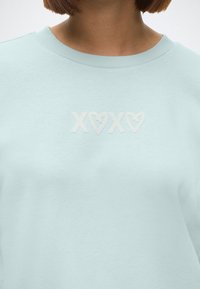 Sweat-shirt bleu clair à col rond avec texte blanc "XOXO" comprenant des formes de cœur sur la partie supérieure de la poitrine et du cou d'une personne.