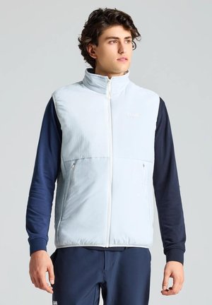 Un homme porte un gilet matelassé blanc à fermeture éclair sur un haut bleu marine à manches longues et un pantalon, devant un fond gris uni.