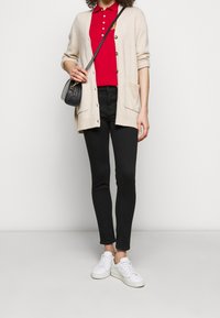 Cardigan en tricot beige avec des boutons, polo rouge, jeans skinny noirs et baskets blanches. Petite sac noir avec bandoulière.