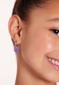 Pendiente con forma circular de carita sonriente morada y herrajes plateados brillantes, llevado en la oreja de un modelo, mostrando detalladas características faciales.
