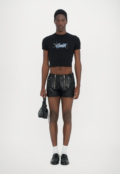 Schwarzes, kurzes T-Shirt mit Grafikdruck, kombiniert mit schwarzen Leder-Shorts mit Reißverschlüssen, weißen Socken und schwarzen flachen Schuhen. Hält eine kleine Tasche.