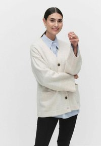 Beige oversized cardigan med V-hals, ribbade ärmslut och två framfickor, dragen över en ljusblå randig skjorta med mörka knappar.