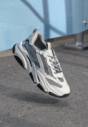 Chunky white and gray Steve Madden sneaker with mesh details hovers above pavement, blue textured wall and shadow in background.