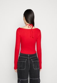 Rød bluse med lange ærmer, bred halsudskæring og bånddetalje bagpå, parret med sorte cargo bukser med flere lommer og hvid syning.