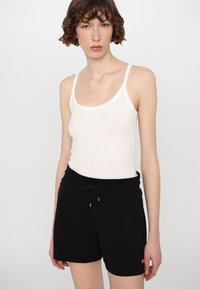 Top blanco sin mangas con tirantes delgados combinado con pantalones cortos negros. Los pantalones cortos tienen una cintura con cordón y un sutil logotipo en el lateral.