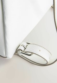 Gros plan d'un sac à main en cuir blanc texturé avec une bandoulière réglable et une boucle argentée sur une surface claire.