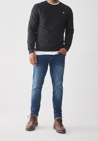 Sudadera negra con cuello redondo y puños de canalé, superpuesta sobre una camisa blanca. Combinada con jeans azules y botas marrones con cordones.
