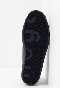 Semelle d'une basket noire avec une surface texturée et un motif de logo partiel audacieux, positionnée debout contre un mur blanc.