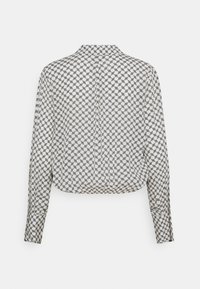 Hvid langærmet bluse med sort geometrisk kædemønster, med knapmanchetter og en lige bagforkant, præsenteret på en hvid baggrund.