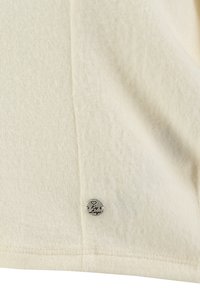 T-shirt lavorato a maglia color crema chiaro con una texture liscia, caratterizzato da un piccolo logo metallico sul bordo. Design semplice senza ulteriori motivi.