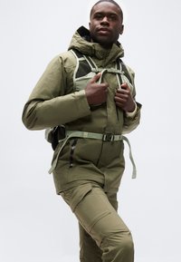 Άνδρας που φοράει χακί εξωτερικό μπουφάν και παντελόνι καμπάνες, ρυθμίζοντας τους ιμάντες ενός χακί σακιδίου πεζοπορίας σε λευκό φόντο.