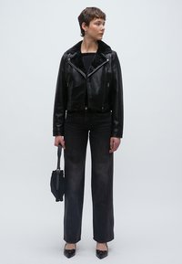 Fekete bőr motorosdzseki műszőrme gallérral, magasderekú fekete farmerrel és fekete, hegyes orrú balerinacipővel párosítva. Egy kis táskát tart a kezében.