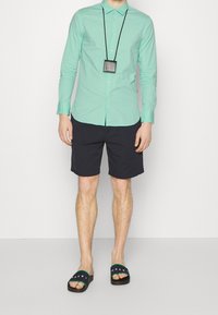 Camicia a maniche corte verde chiaro con bottoni, pantaloni corti blu navy e sandali neri con dettagli verdi. Indossa una collana con un pendente rettangolare.