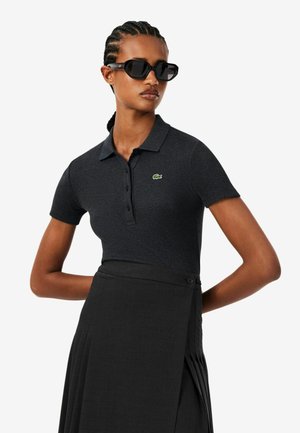 Femme aux cheveux tressés portant des lunettes de soleil noires, un polo sombre à manches courtes avec un petit logo, et une jupe plissée taille haute.