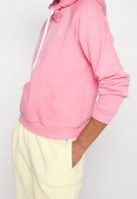 Rosa sweatshirt med huva, vit dragsko, frontficka, ribbade ärmslut och nederkant, tillverkad av mjukt tyg, parat med ljusgula byxor.