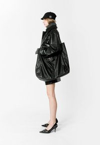 Abrigo oversized de cuero negro con textura suave; combinado con una gorra negra, bufanda gris y tacones negros brillantes con adornos florales.