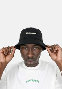 Cappello a secchiello nero con il logo "SIXTHUNE" in argento sulla parte anteriore, indossato da una persona. T-shirt bianca con testo verde visibile sotto.