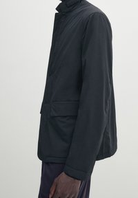 Giacca nera, leggera, con colletto alto, chiusura a bottone e tasche laterali. Presenta una texture liscia e un design minimalista.