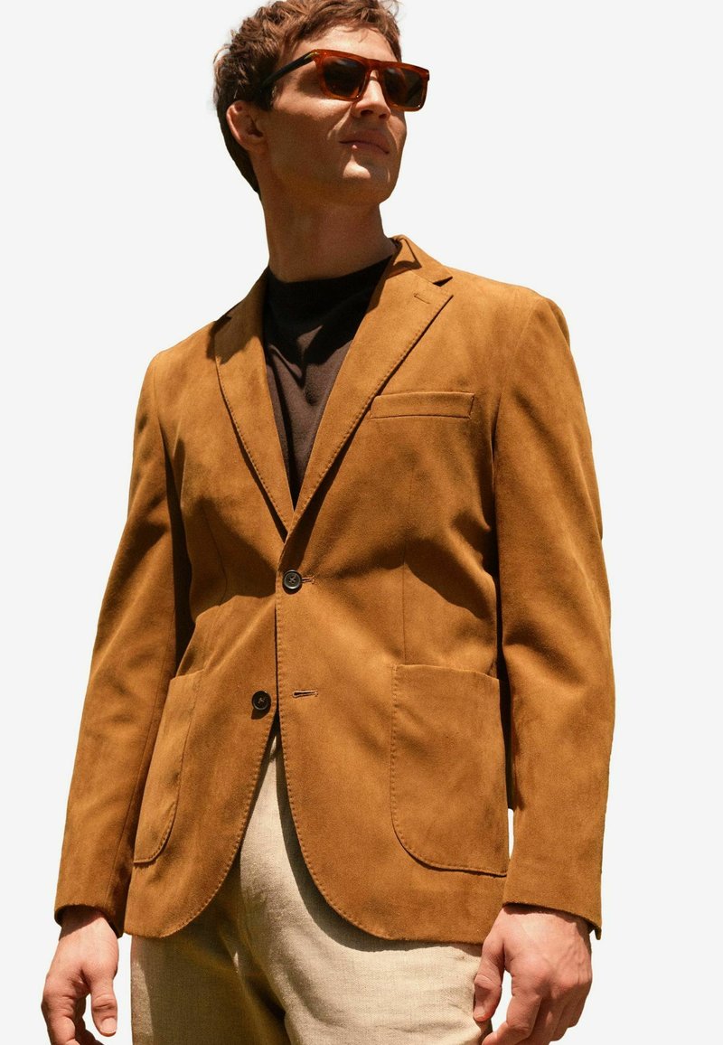 Barna szarvasbőr blézer bevágott hajtókával, két elülső zsebbel és két gombbal. Sötét felsőre véve, világos színű nadrággal párosítva.