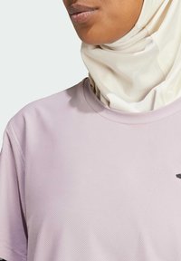 Ljuslila, texturerad träningsskjorta med rund halsringning och en liten svart logotyp. En krämfärgad huvudduk bärs under.